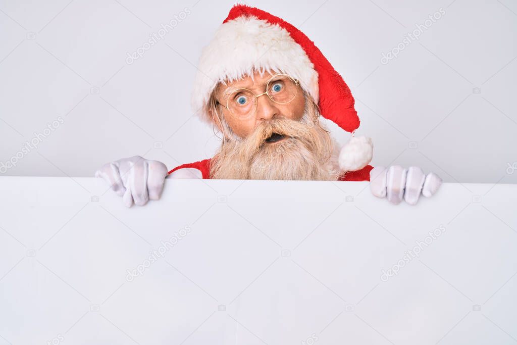 Old senior man with grey hair and long beard wearing santa claus costume holding banner in shock face, looking skeptical and sarcastic, surprised with open mouth 