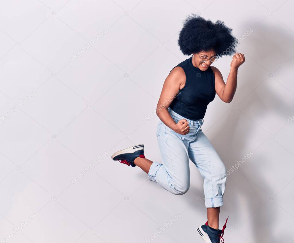 Young beautiful african american woman wearing casual clothes and glasses smiling happy. Jumping with smile on face doing winner gesture with fists up over isolated white background