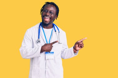 Doktor steteskop ve kimlik kartı takmış, gülümseyen ve kameraya bakıp iki eli ve parmağı yandan gösteren Afrikalı genç bir Amerikalı.. 