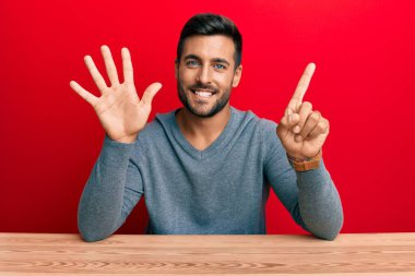 Yakışıklı İspanyol bir adam sıradan giyinmiş masanın üzerinde oturuyor ve 6 numaralı parmaklarıyla gülümsüyor kendinden emin ve mutlu.. 