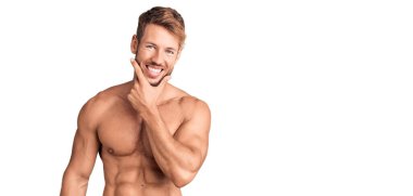 Genç, beyaz, üstsüz bir adam kameraya bakıyor. Kollarını kavuşturup gülümsüyor ve çenesini kaldırıyor. Olumlu düşünmek. 