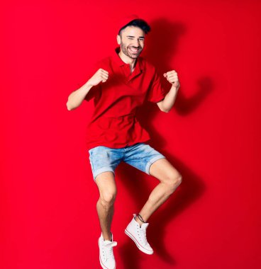 Şapkalı genç teslimatçı mutlu gülümsüyor. Yüzünde tebessümle zıplıyor, kırmızı arka planda yumruklarıyla kazanan jestler yapıyor.