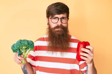 Uzun sakallı, elinde brokoli ve kırmızı biber tutan yakışıklı kızıl saçlı adam yüzünde ciddi bir ifadeyle rahatlamış. Basit ve doğal bir şekilde kameraya bakıyorsun.. 