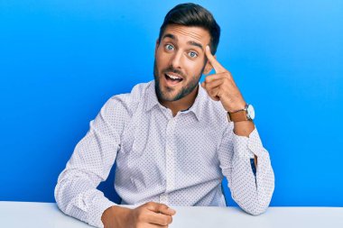 İş kıyafetleri giymiş yakışıklı İspanyol bir adam masaya oturmuş gülümsüyor tek parmağıyla işaret ediyor, harika bir fikir ya da düşünce, iyi bir hafıza. 