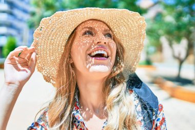 Yaz şapkası takmış, mutlu bir şekilde gülümseyen güzel sarışın bir kadın. Şehrin caddesinde yüzünde gülümsemeyle dikilirken.