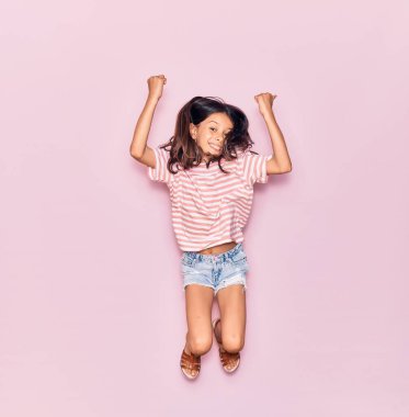 Sevimli İspanyol kız günlük kıyafetler giyip mutlu gülümsüyor. İzole edilmiş pembe arka planda yüzünde gülümsemeyle zıplıyorsun.