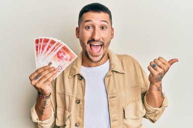 Dövmeleri olan yakışıklı bir adam elinde 20 İsrail şekeri banknotları başparmağıyla yan tarafa bakıyor mutlu bir şekilde ağzı açık gülümsüyor. 