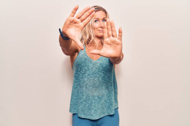 Orta yaşlı beyaz kadın spor giysisi giyiyor. El ve parmaklarıyla çerçeve yapıyor. Kamera açısı var. 