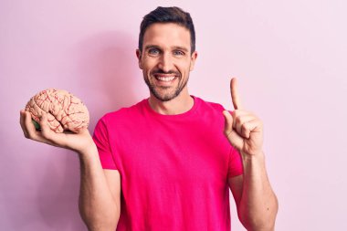 Genç yakışıklı adam, beyni soyutlanmış pembe arka planda tutarak mutlu bir yüzü işaret eden bir fikir veya soruyla gülümsüyor.