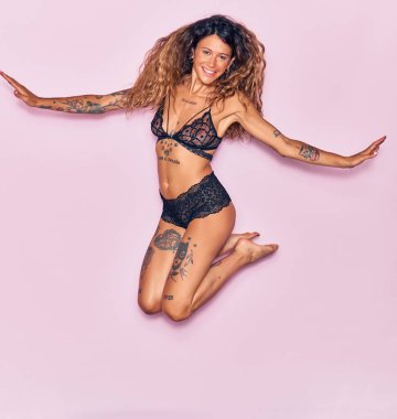 Genç, güzel, kıvırcık, dövmeli, iç çamaşırı giyen mutlu bir kadın. Yüzünde ve kollarında gülücüklerle pembe bir arka planda atlamak.