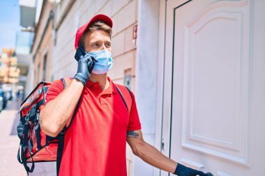 Beyaz postacı kırmızı üniforma ve teslimat çantası giyiyor ve akıllı telefon kullanarak koronavirüs güvenlik maskesi takıyor.