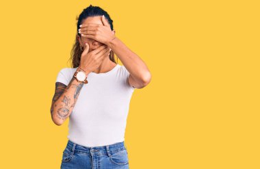 Dövmeli, beyaz tişörtlü, gözleri ve ağzı elleri bağlı, şaşırmış ve şok olmuş genç bir İspanyol kadın. duygu gizleme 