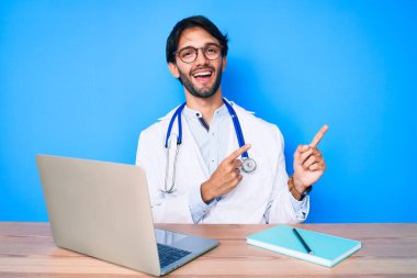 Yakışıklı İspanyol üniformalı doktor klinikte çalışıyor gülümsüyor ve kameraya bakıyor iki eliyle ve parmaklarıyla yandan işaret ediyor.. 