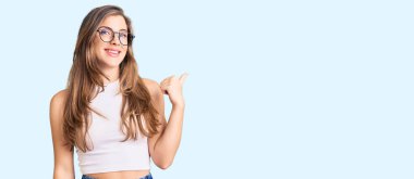 Gündelik kıyafetler ve gözlüklerle gülümseyen, gülümseyen ve baş parmağıyla yüzünü gösteren güzel beyaz bir kadın.. 