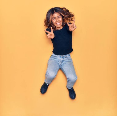 Mutlu gülümseyen genç, güzel bir Latin kadın. Yüzünde gülümsemeyle zıplıyor zafer işareti yapıyor izole edilmiş sarı arka planda