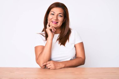 Orta yaşlı esmer, Latin bir kadın. Masada oturmuş gülümsüyor ve elleri ve çenesi çaprazlanmış kameraya bakıyor. Olumlu düşünmek. 