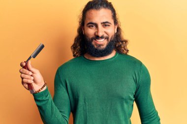 Genç Arap adam berber jiletini tutuyor. Olumlu ve mutlu görünüyor. Duruyor ve gülümsüyor. 