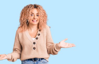 Kış kazağı giyen, kıvırcık saçlı, kafası karışık ve kolları ve elleri havada genç, sarışın bir kadın. şüphe kavramı. 