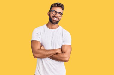 Gündelik kıyafetler ve gözlük takan genç İspanyol adam kameraya bakarken kollarını kavuşturup gülümsüyor. pozitif kişi. 