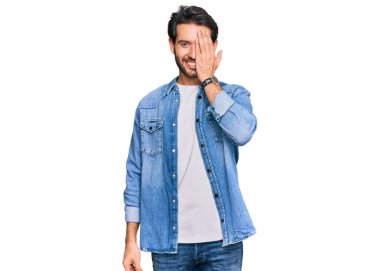 Latin asıllı genç adam gündelik giysiler giyiyor. Bir gözünü eliyle kapatıyor. Yüzünde kendine güvenen bir gülümseme ve sürpriz bir duygu var.. 