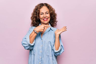Orta yaşlı güzel bir kadın pembe arka plan üzerinde sıradan bir kot gömlek giyiyor hayret ediyor ve kameraya gülümsüyor, elini uzatıyor ve parmağıyla işaret ediyor..