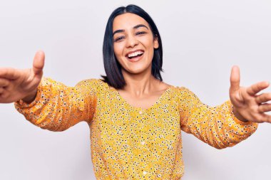 Gündelik giysiler giymiş genç ve güzel bir Latin kadın kameraya bakıp sarılmak için kollarını açmış gülümsüyor. Mutluluğu kucaklayan neşeli bir ifade.. 