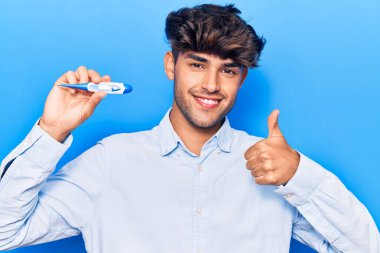 Genç İspanyol adam termometreyi tutuyor mutlu ve pozitif gülümsüyor, başparmağıyla mükemmel ve onay işareti yapıyor. 