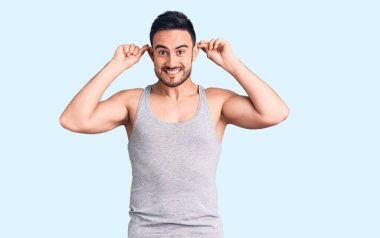 Mayo ve kolsuz tişört giyen genç yakışıklı adam gülümsüyor kulakları parmaklarıyla çekiyor, komik bir jest. deneme sorunu 