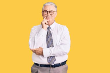 İş kıyafetleri giymiş yaşlı gri saçlı adam kameraya kendinden emin bakıyor kollarını kavuşturup gülümseyerek ve çenesini kaldırarak. Olumlu düşünmek. 