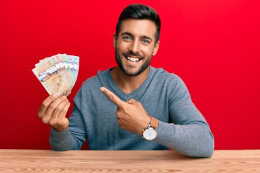 Kanada doları tutan yakışıklı İspanyol adam mutlu bir şekilde elini ve parmağını gösterdi. 