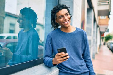Genç Afrikalı Amerikalı mutlu gülümsüyor. Duvara dayanmış akıllı telefon kullanıyor..