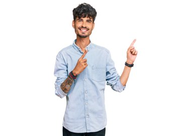 Gündelik kıyafetler giyen genç İspanyol adam gülümseyip kameraya bakıyor ve iki eliyle ve parmaklarıyla kenarı işaret ediyor.. 