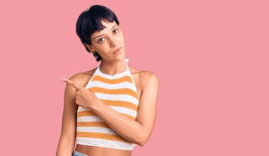 Kısa saçlı genç esmer kadın, günlük kıyafetler giyiyor. El parmağı yanda, ciddi ve sakin bir yüz ifadesi sergiliyor. 