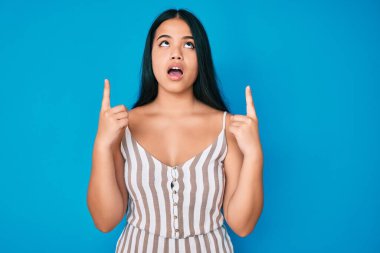 Genç ve güzel Asyalı kız gündelik giysiler giyip şaşkın şaşkın yukarı bakıyor ve parmaklarıyla işaret ediyor ve kollarını kaldırıyor.. 