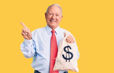 Yakışıklı, beyaz saçlı, şık kıyafetler giyen, para çantası tutan, gülümseyen, elini ve parmağını yana doğru gösteren bir adam. 