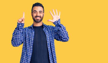 Latin asıllı genç adam gündelik giysiler giyip altı numaralı parmaklarıyla işaret ederken kendinden emin ve mutlu bir şekilde gülümsüyor.. 