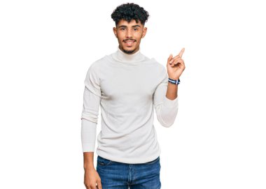 Genç Arap adam, sıradan bir kazak giyiyor. Yüzünde kocaman bir gülümseme var. Parmağıyla kameraya bakıyor.. 