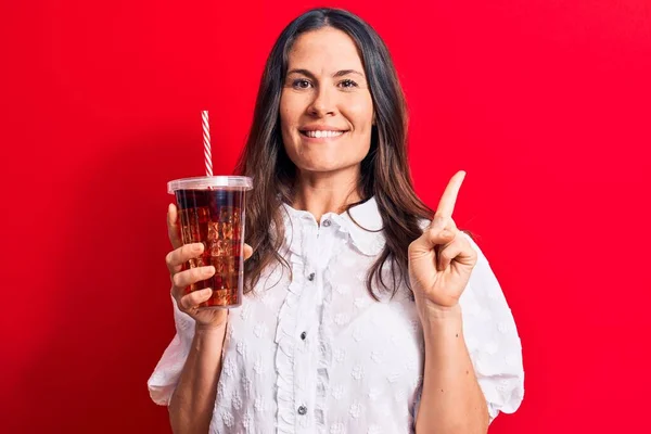 美丽的黑发女人喝可乐提神饮料时 用稻草盖在红色的背景上 开心地微笑着 手指手画脚地指向旁边 — 图库照片