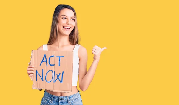 Young beautiful girl holding act now banner pointing thumb up to the side smiling happy with open mouth