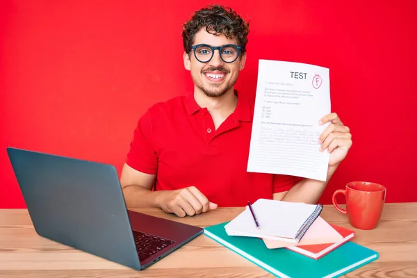 Young caucasian man with curly hair sitting on the table showing failed exam looking positive and happy standing and smiling with a confident smile showing teeth