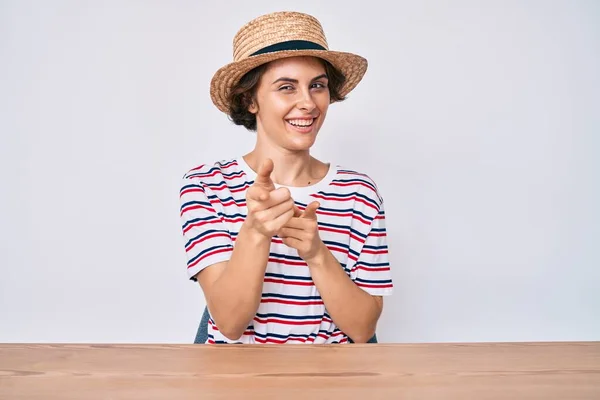 Young hispanic woman wearing casual clothes and hat sitting on the table pointing fingers to camera with happy and funny face. good energy and vibes.