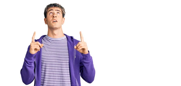 年轻英俊的男子穿着休闲的紫色运动衫 惊讶地抬起头 用手指和胳膊指指着 — 图库照片
