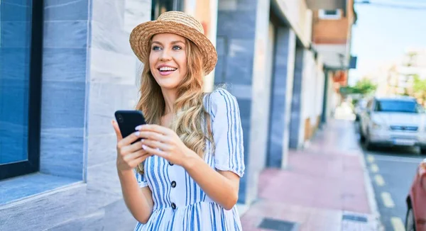 年轻美丽的金发女子戴着夏帽在度假时快乐地笑着 在城市街道上用智能手机笑着站在那里 — 图库照片