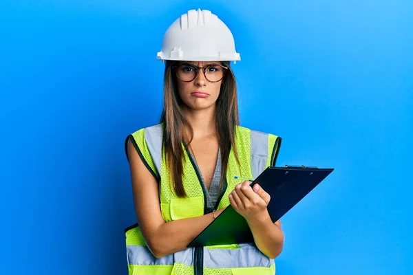 Young hispanic woman wearing safety helmet holding clipboard skeptic and nervous, frowning upset because of problem. negative person.