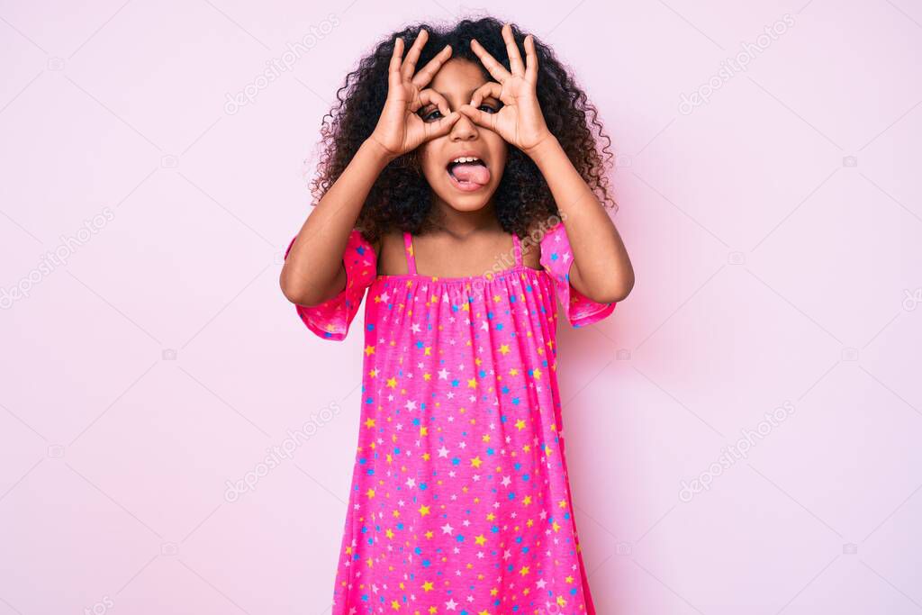 African american child with curly hair wearing casual dress doing ok gesture like binoculars sticking tongue out, eyes looking through fingers. crazy expression. 