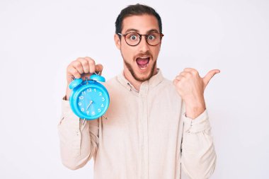 Çalar saati tutan yakışıklı genç bir adam başparmağını yana doğru uzatıyor ve ağzı açık gülümsüyor. 