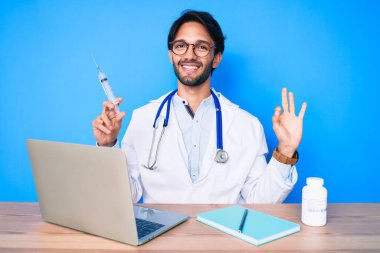 Klinikteki yakışıklı İspanyol doktor elinde şırıngayla tamam işareti yapıyor, gülümsüyor ve mükemmel bir sembol oluşturuyor. 