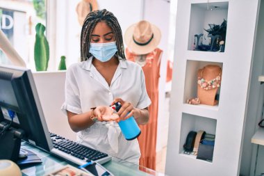 Genç Afrikalı Amerikalı kadın gülümseyerek kasada çalışırken Coronavirus güvenlik maskesi takıyor ve perakende dükkanında dezenfektan jeli kullanıyor.