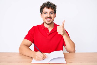 Kıvırcık saçlı genç beyaz adam masaya oturmuş gülümseyen ve olumlu gülümseyen bir not defteri yazıyor, başparmağı mükemmel ve onay işareti yapıyor. 