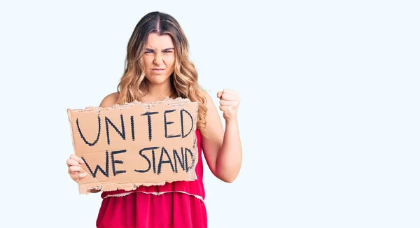Young caucasian woman holding united we stand banner annoyed and frustrated shouting with anger, yelling crazy with anger and hand raised